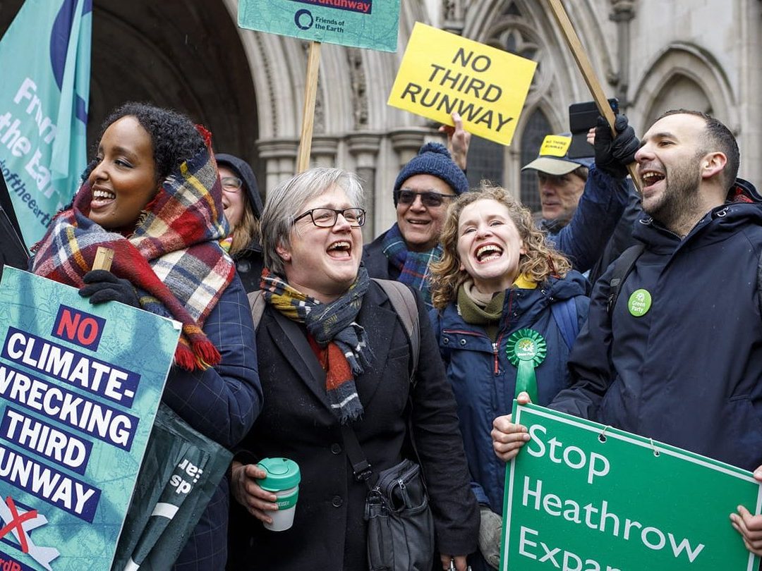 Zack Polanski protesting the heathrow expansion in crowd with fellow green party members. The crowd appears to be cheering and smiling, holding colourful placards.